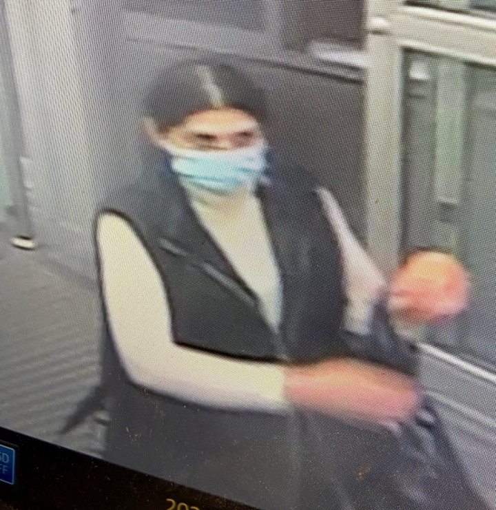 A woman is pictured exiting a store. She has long dark hair parted in the middle of her head with gray roots exposed. Her hair is pulled back in a pony tail. She is wearing a blue face mask, a white long sleeved turtleneck shirt with a black vest on top. She is carrying a large black bag.