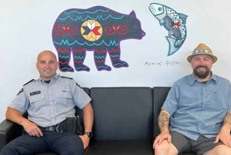 A uniformed RCMP Officer sits on a black couch, next to man wearing a hat. Both men are smiling. They sit in front of a large, painted mural featuring a large black bear, a fish, an eight-pointed 'Mi'kmaq Star' and artistic renderings of Mi'kmaq applique patterns and imagery.
