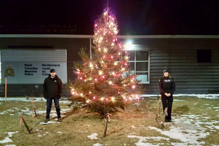 Un homme portant un chapeau noir, un manteau noir et un jean se tient à côté d'un arbre de Noël illuminé. Une agente de la GRC en uniforme se tient de l'autre côté de l'arbre. La photo a été prise le soir en face d'un détachement de la GRC.