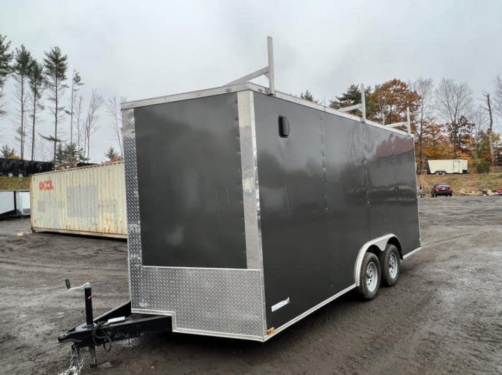 A dark grey colored enclosed utility trailer is parked on a gravel lot. A Sea-Can storage container and a vehicle is observed in the background.