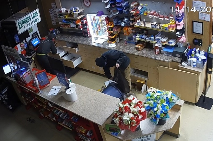 Two masked individuals are inside a store, after hours. One is opening drawers behind the cash while the other is placing merchandise into a bag.
