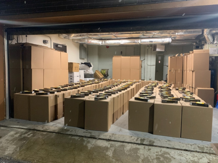 A garage door is open. Inside a large number of large cardboard boxes are displayed with a carton of tobacco placed on the top of each box.