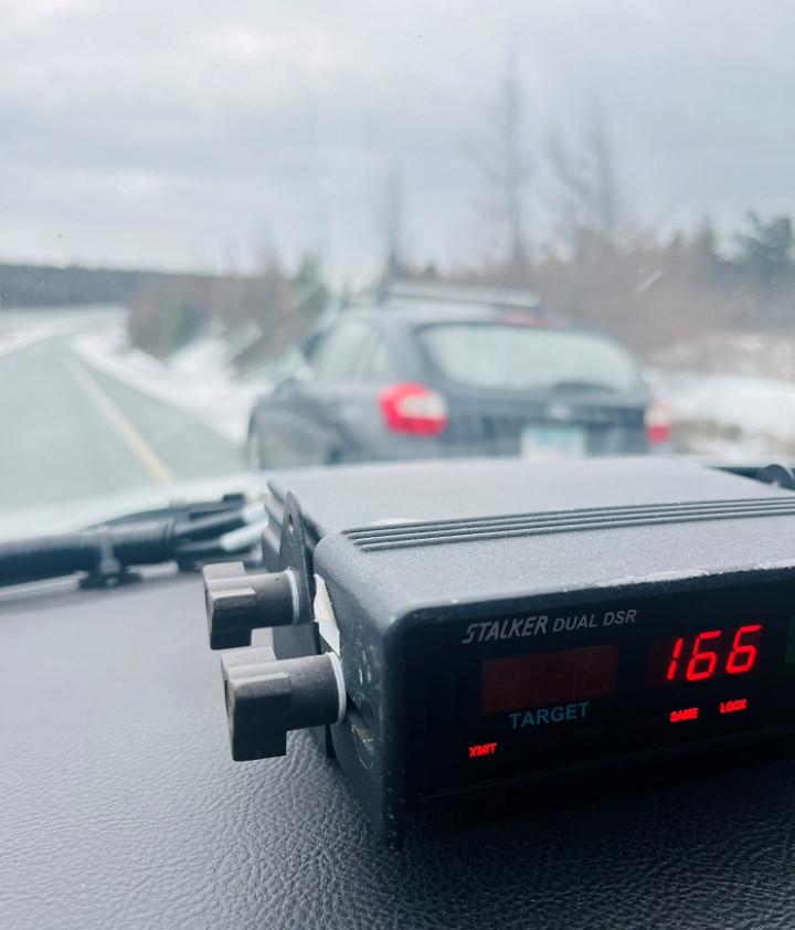 A picture is taken from inside a police vehicle. A radar unit is sitting on the dash displaying the number 166. A car is pulled over on the side of the road in front of the police vehicle on an overcast winter day. 