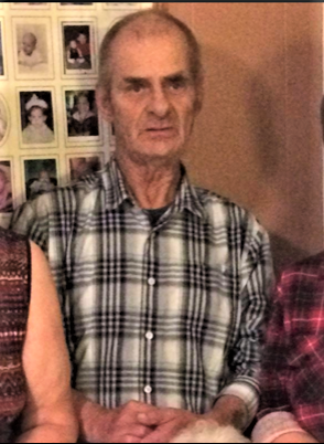 An elderly man wearing a plaid button up shirt is pictured seated from the waist up. He is balding with dark grey hair and is clean shaven.