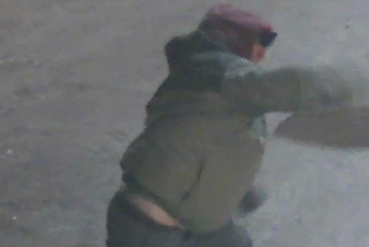 An individual, who appears to be a man, is pictured wearing black boots, jeans and a green colored jacket, with a burgundy hood pulled up overhead. The individual is throwing something.