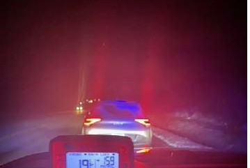 Un radar à l'intérieur d'une voiture de police affiche une vitesse de 159 km/h. Une voiture est garée devant la voiture de police sur le bord d'une autoroute, en soirée.