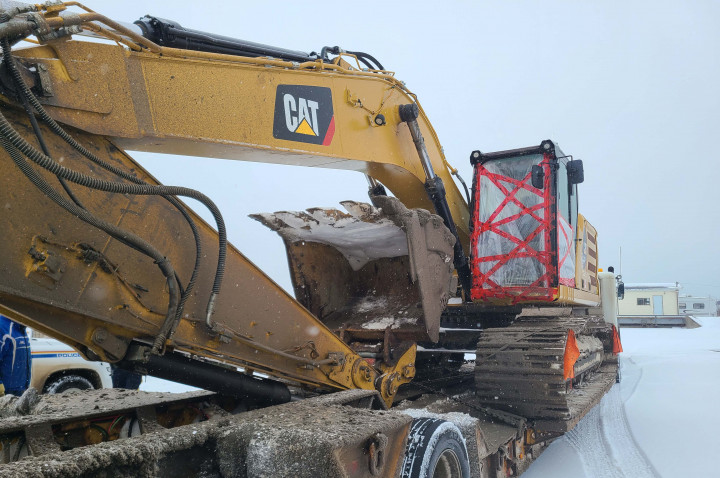 Une excavatrice dont la porte vitrée de la cabine est cassée se trouve sur une remorque à plate-forme.