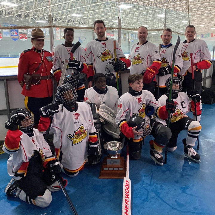 The RCMP Christie Lake Kids hockey team, along with S/Sgt. Boissonneault who is in red serge uniform, pose with the STAR Cup. Team members are standing and kneeling off the ice in front of a rink.