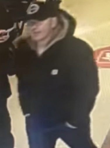 A Caucasian man is pictured wearing a black ball cap and a black hooded winter coat inside a store.