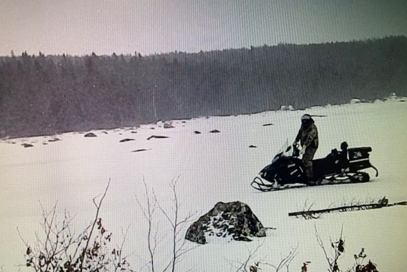 Image en noir et blanc d'une personne en motoneige sur un terrain dégagé et enneigé. Des arbres sont visibles à l'arrière-plan.