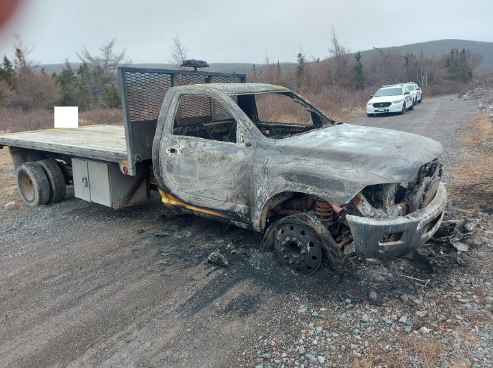 A pickup truck with a flat bed is extensively damaged by fire and is parked on a gravel surface. Two RCMP police vehicles are parked along a gravel road behind the truck.