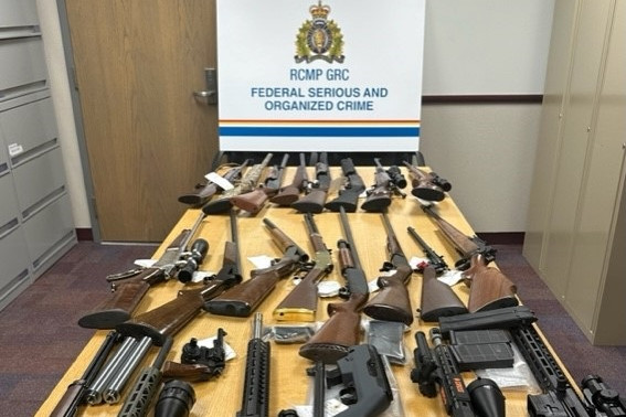 Twenty-five various types of firearms are displayed on a wooden table. A sign at the end of the table reads 'RCMP GRC Federal Serious and Organized Crime' (Photo: RCMP NL)