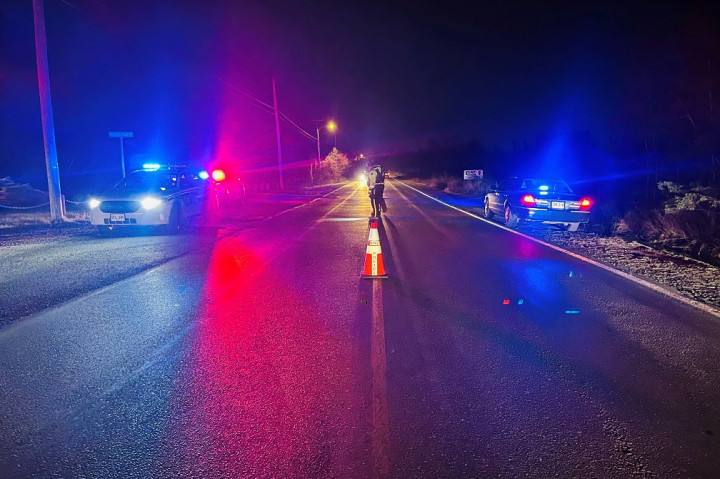 Two police vehicles with emergency lights activated are parked on opposite sides of a roadway at night. Two RCMP officers are stood on the roadway behind a pilon.