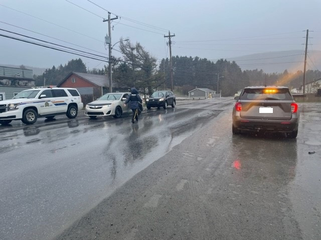 A RCMP officer is walking down a roadway towards two vehicles parked behind a marked police vehicle on a rainy day.
