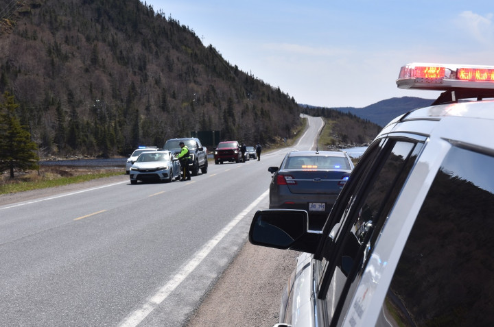 Two police vehicles are pulled over on the side of a highway. In the opposite lane of travel, three police officers are standing on the roadway with four vehicles stopped during a daytime checkpoint.