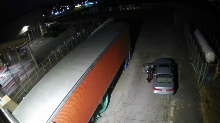 A surveillance image of an individual standing beside the driver side of a grey car at night.