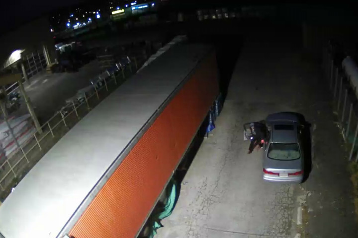 A surveillance image of an individual exiting the driver side of a grey car at night.