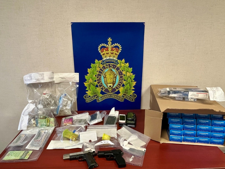 A number of items seized by RCMP are displayed on a table, including quantities of suspected cocaine, illicit cannabis, contraband tobacco, cash, two hand guns (pellet guns) and other evidence consistent with drug trafficking.