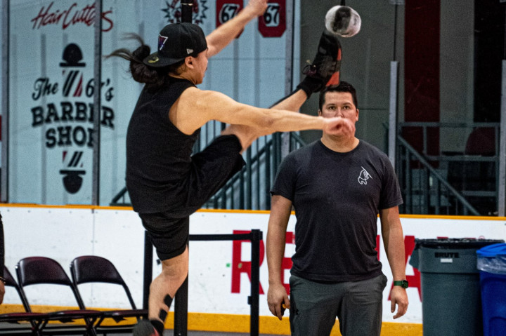 Inuit games, one-foot high kick competition. In an arena, a person is jumping and simultaneously kicking one-leg up to touch a ball that is suspended high in the air. Another person is standing to supervise the competitor. 