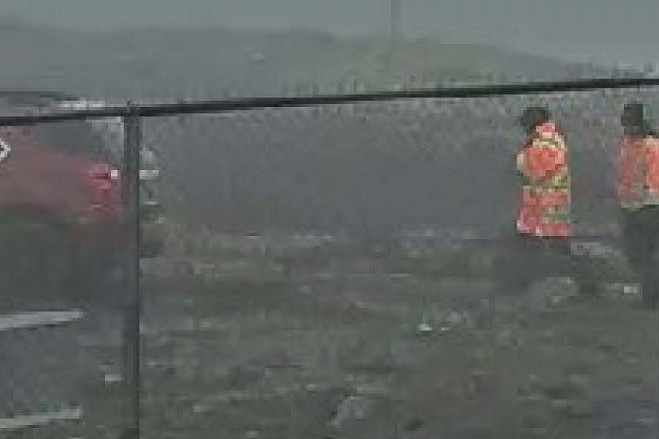 A surveillance image shows two individuals wearing high-visibility jackets walking towards a red hatch-back vehicle parked outside a fenced area.
