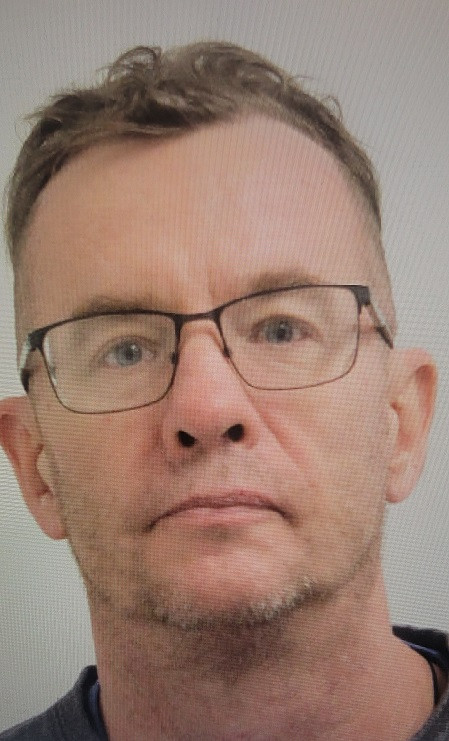A man is pictured from the neck up. He is wearing eye glasses and has dark blonde hair.