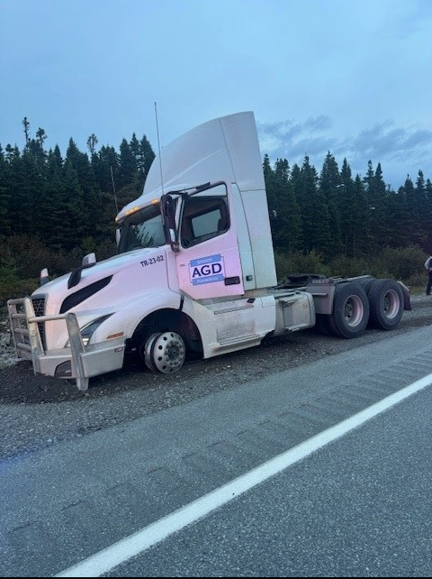 Un camion de transport dans l'accotement en gravier d'une route. Le devant du véhicule est enfoncé dans le gravier et le pneu avant côté conducteur est dégonflé.