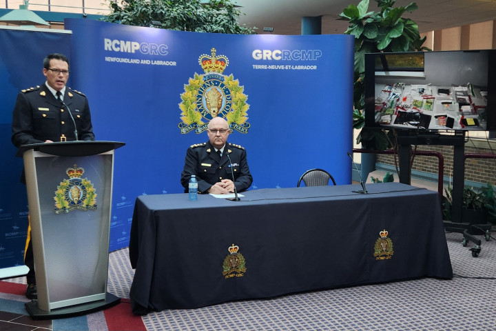 A uniformed police officer is standing behind a podium. Another uniformed police officer sits at a table beside a TV displaying the various items from a drug seizure. A blue RCMP banner with the RCMP crest can be seen in the background.
