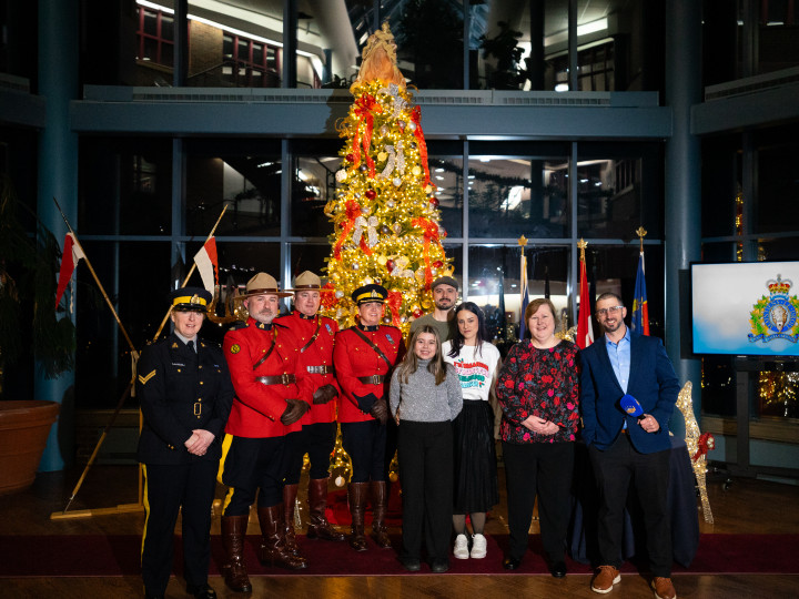 Uniformed RCMP officers, a girl and her parents, the weatherman news anchor, and a woman stand in front of a lit Christmas tree. 