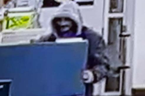 A man wearing a dark jacket, a grey hood over his head, and dark facial hair standing behind a store display.