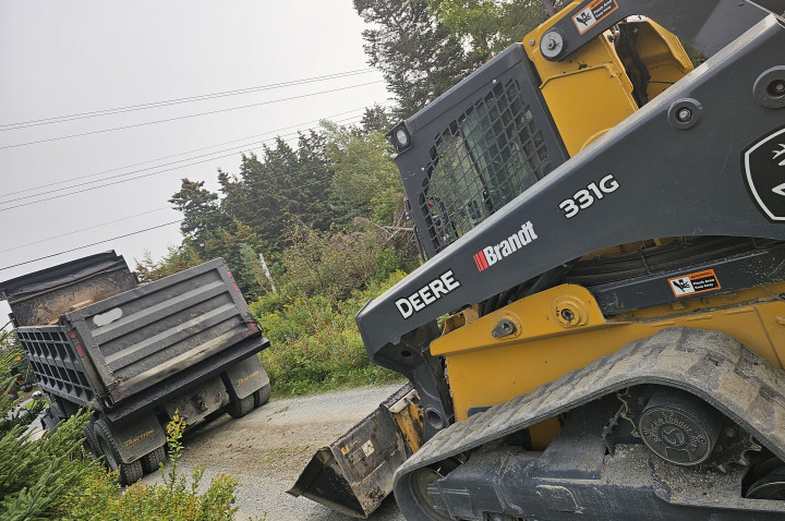 A yellow John Deere skid-steer loader behind a dump truck on a dirt road.