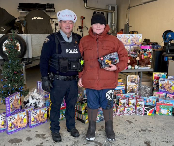 A uniformed RCMP officer wearing a Santa hat is standing next to a youth who is holding a toy. There are toys stacked behind them both on a table and on the floor. 