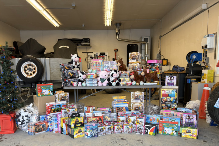 Various unwrapped toys are stacked on a floor and on a table inside of a garage. There is a Christmas tree next to the table. 