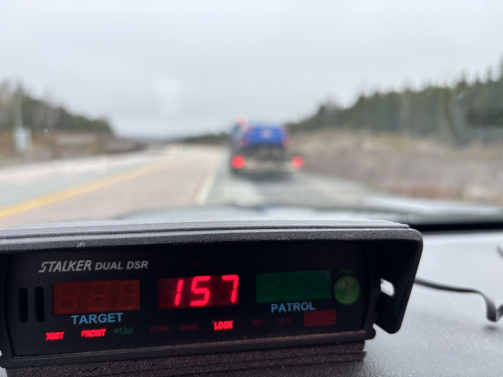A car is pulled over on the side of a road. A police vehicle's radar is pictured in the foreground displaying a speed of 157 km/h. 