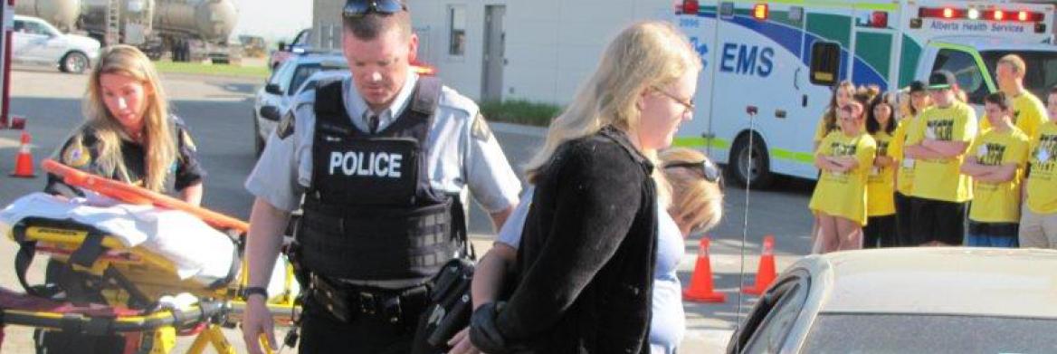 Students at a mock car crash watch a female police officer handcuff a woman next to a car. A male police officer and a paramedic are also on scene.