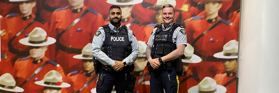 Two male RCMP officers stand side-by-side smiling in front of a mural of Mounties in red serge.