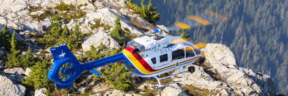 Un hélicoptère affichant le mot police survole la cime rocheuse d'une montagne surplombant une vaste forêt.