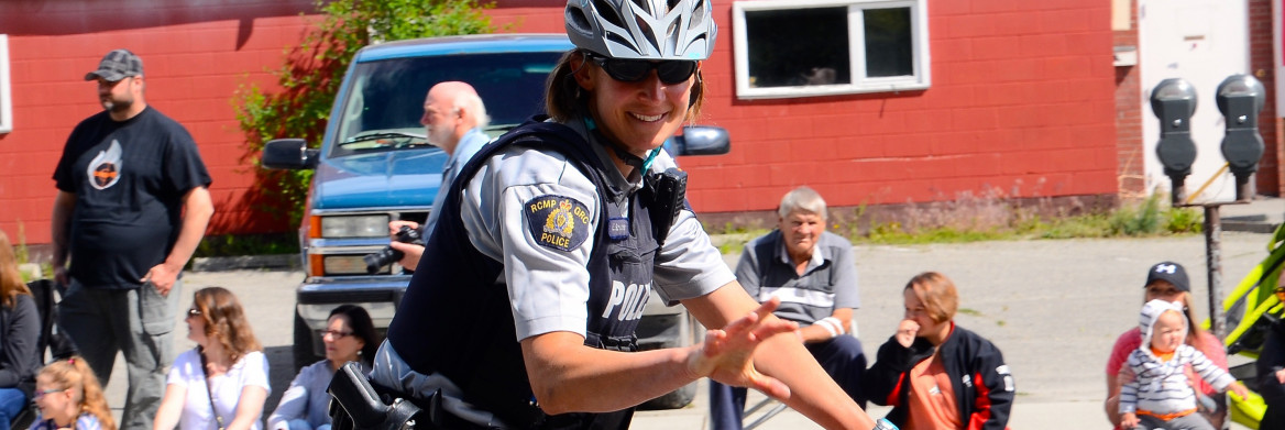 On a sunny day, a female RCMP officer waves to a small crowd of people as she rides past them on a police-issued bicycle wearing sunglasses and a grey helmet.