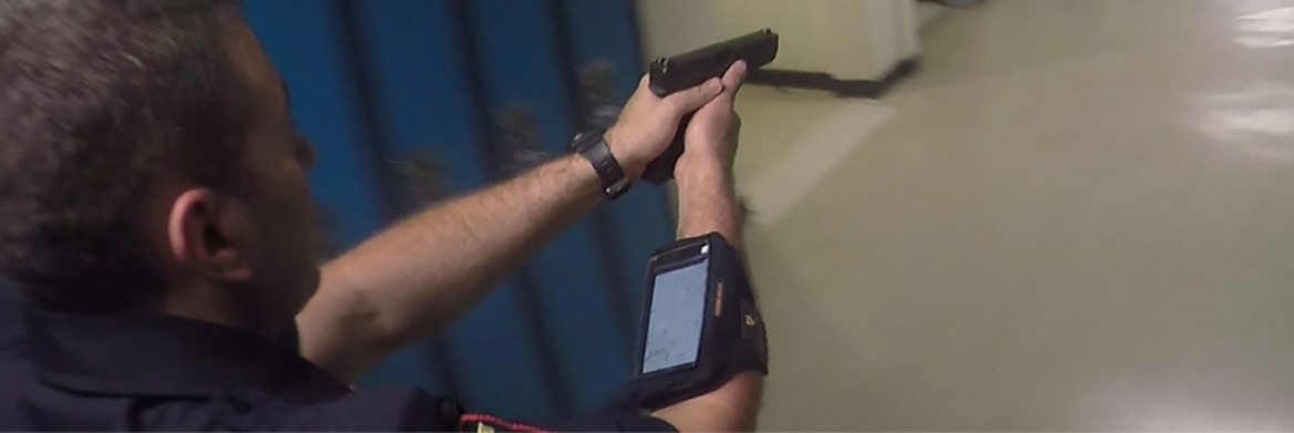 Policier dans un corridor, vu de dos, braquant son arme et portant un cellulaire fixé à l'avant bras.