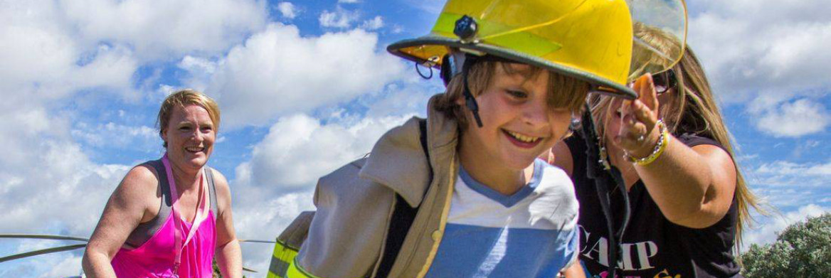 A young boy wearing a firefighter's jacket and helmet runs and smiles. A woman stands behind him smiling.