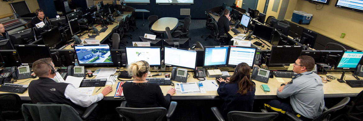 A male RCMP officer pictured at bottom right sits in a room with full of computer screens and other RCMP employees.