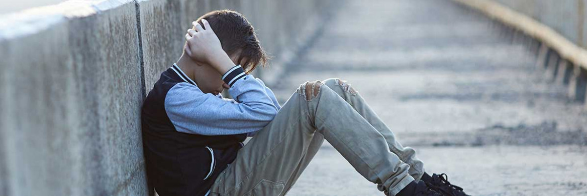 A boy with his head is his hands sitting on a concrete sidewalk.