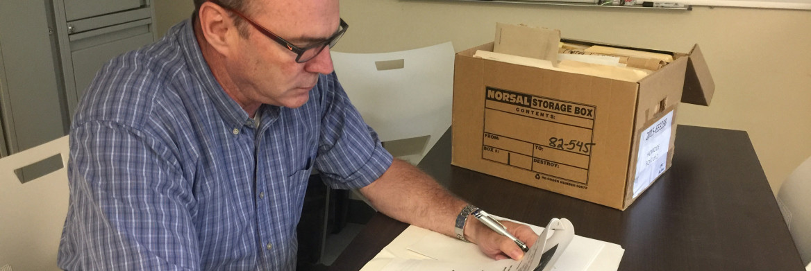 A male RCMP officer in civilian clothes looks over some papers, with a cardboard box on the table next to him.