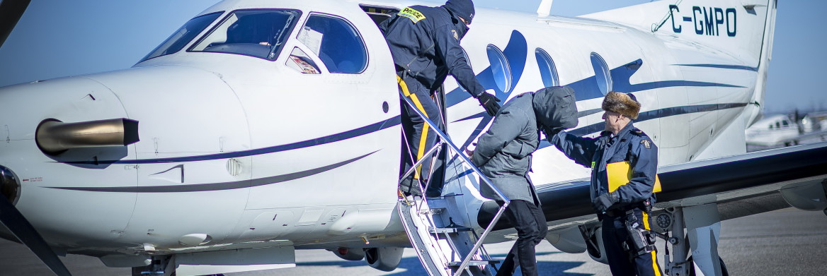 Two male RCMP officers escort a handcuffed individual with hood over his face away from an airplane.