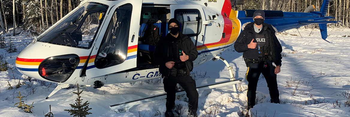 Devant un hélicoptère, un policier de la GRC et un agent de conservation se tiennent dans la neige profonde, le pouce levé. 