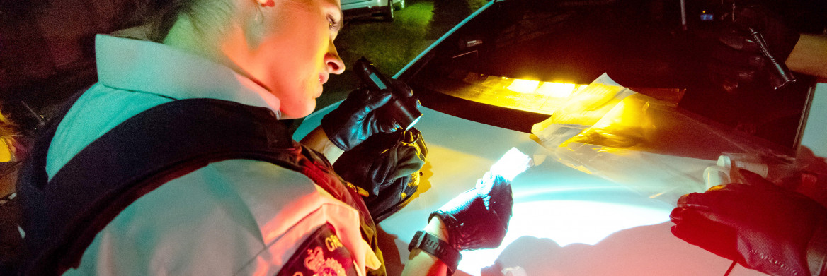 A female RCMP officer uses a flashlight to inspect a small package that she is holding with gloved hands. It is night and she is lit by the lights of a police car. 