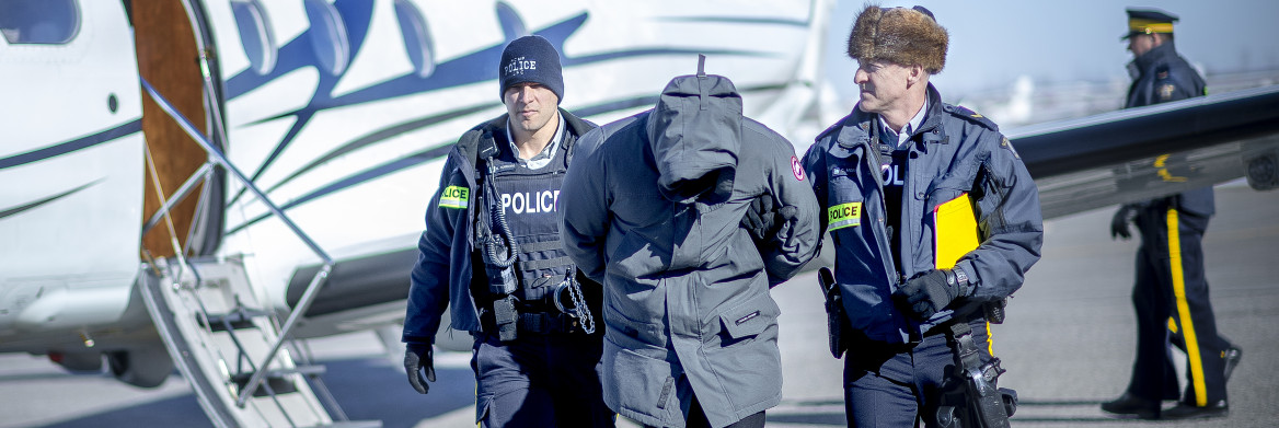Deux agents de la GRC escortent un suspect menotté sur une piste d'aéroport avec un avion garé en arrière-plan.