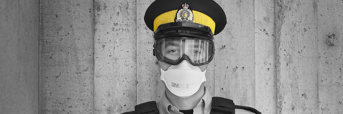 Un policier en uniforme portant un masque blanc ajusté et des lunettes de protection. Il est debout dehors devant un mur gris.