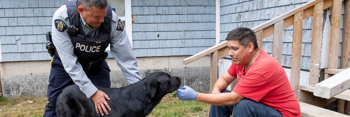 An Indigenous male RCMP officer leans down to pet a black dog. An Indigenous man in a red shirt sits on wooden stairs next to them.