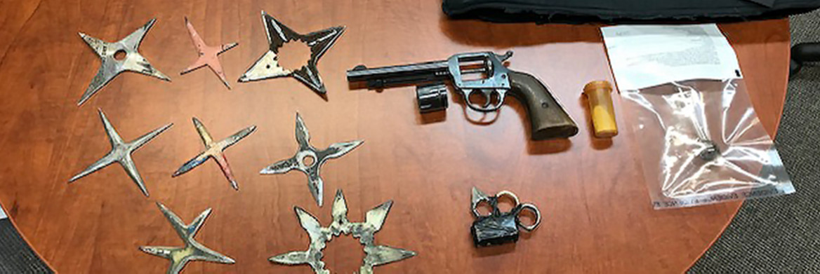 A gun, several star-shaped blade weapons, and a capsule of drugs are displayed on a table. 