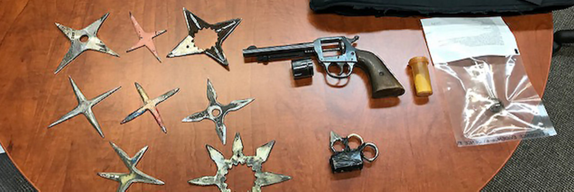 A gun, several star-shaped blade weapons, and a capsule of drugs are displayed on a table.